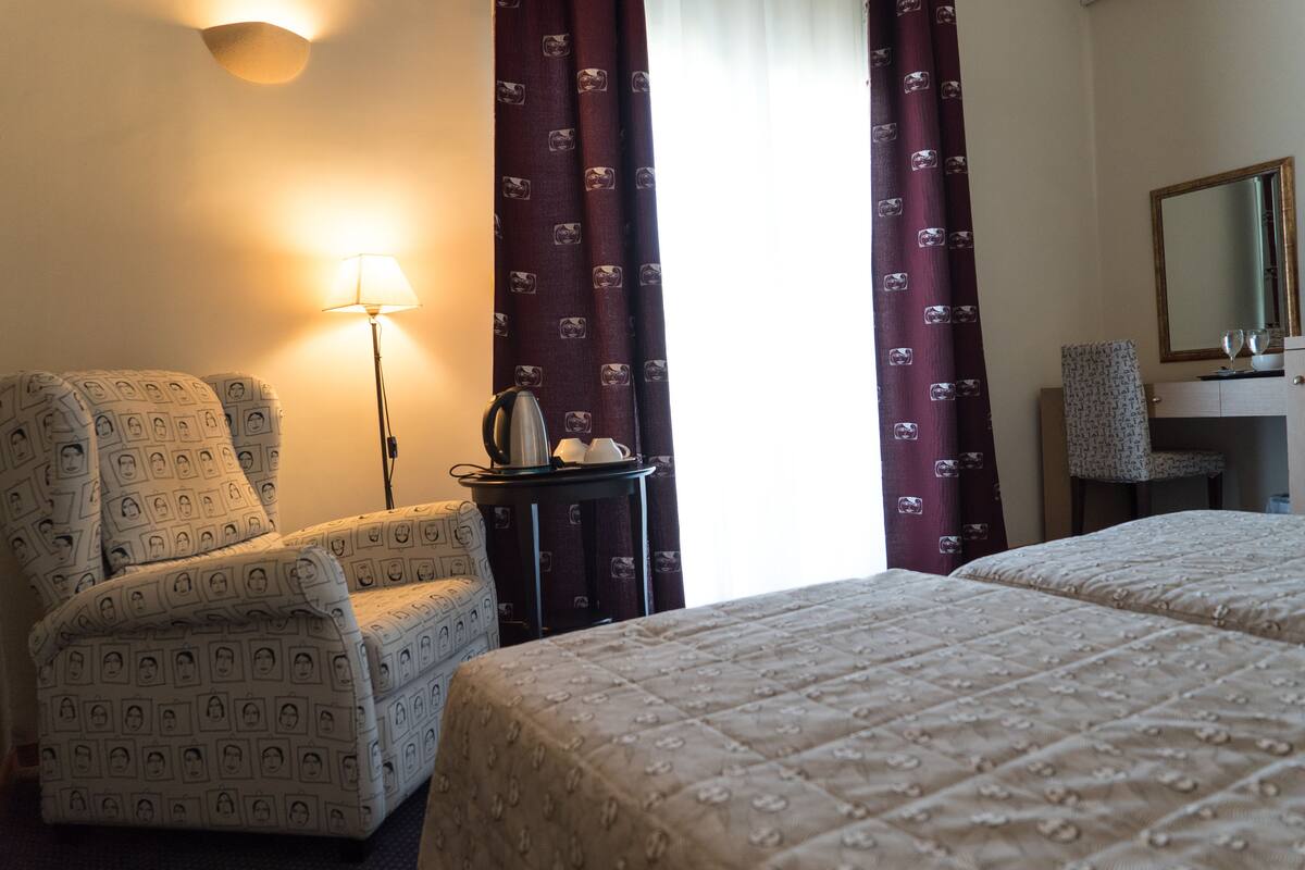 A hotel room with two beds, a chair, a lamp, a small table with a kettle, and a mirror.