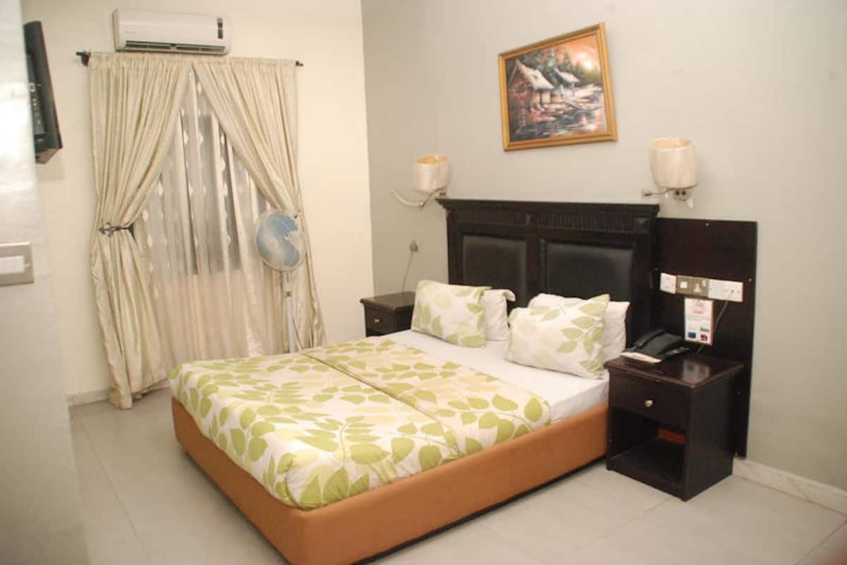 A hotel room with a bed, a bedside table, a nightstand, an air conditioner, a wall-mounted fan, a painting, and a curtain.