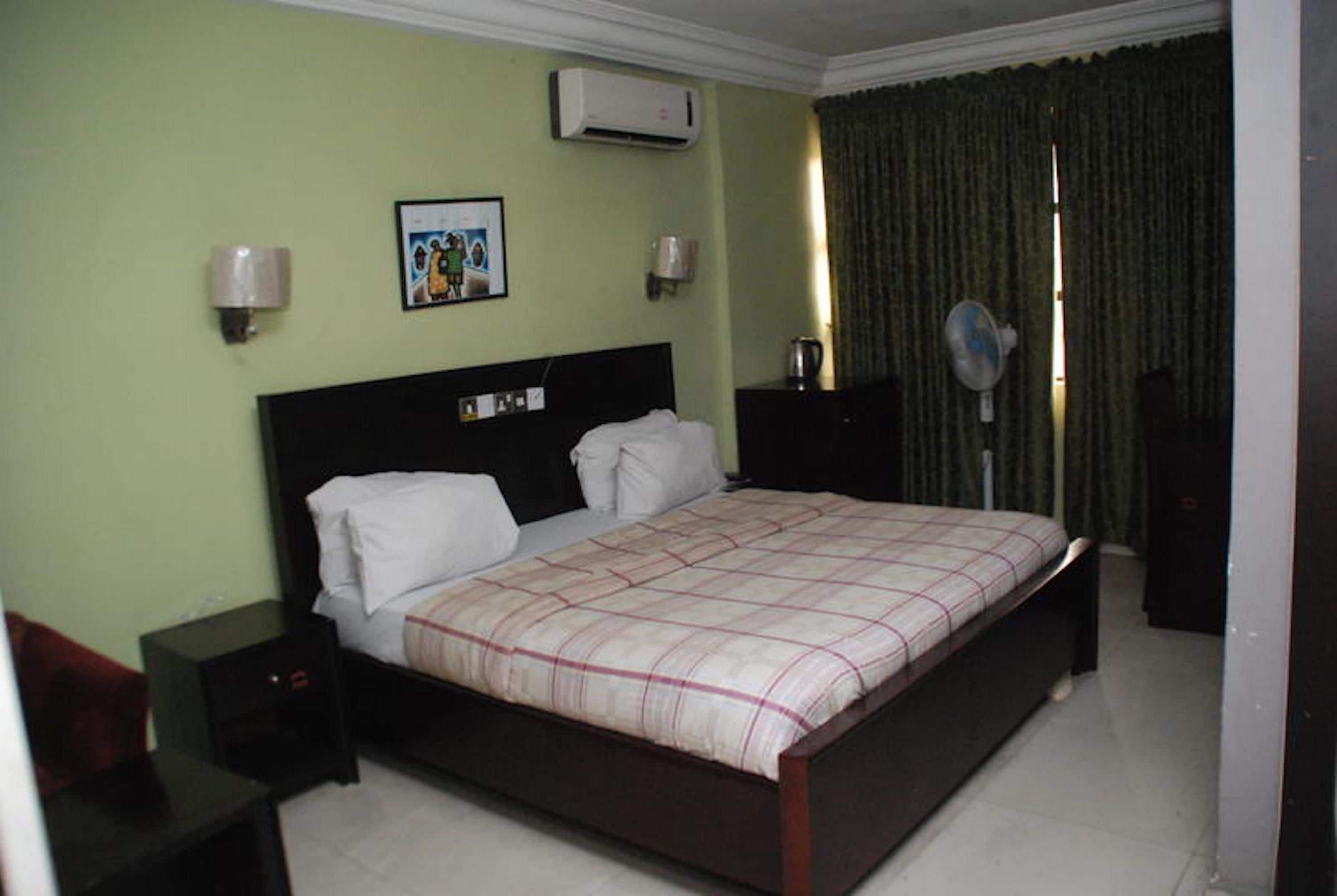 A hotel room with a bed, bedside tables, an air conditioner, a wall-mounted fan, and a curtain.