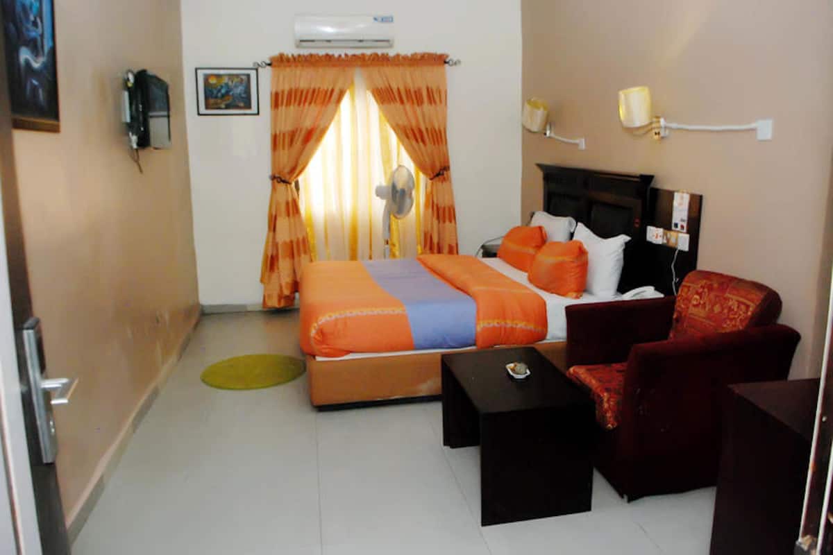 A hotel room with a bed, a red armchair, a small table, a fan, and a wall-mounted air conditioner.