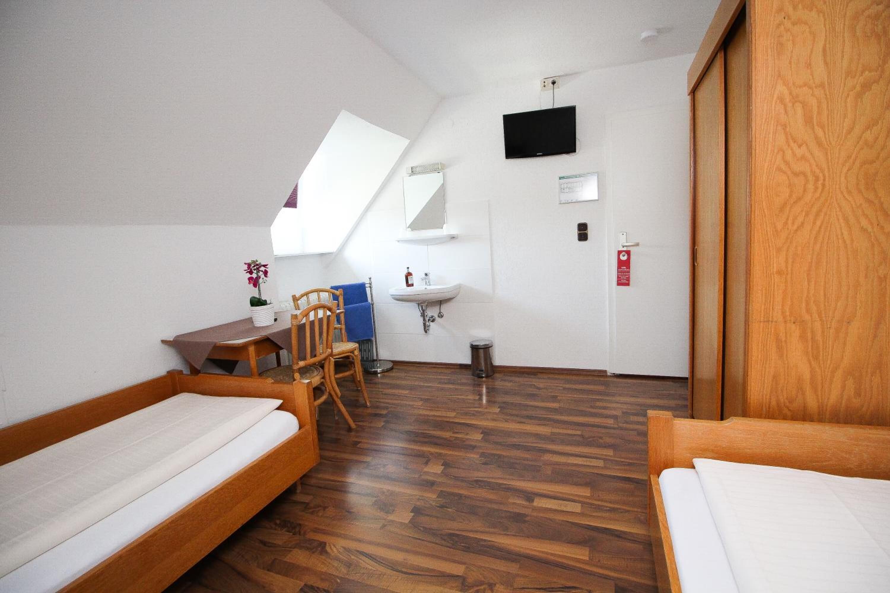 Una habitación pequeña con cama de madera, un escritorio con silla, un lavamanos, un espejo y un televisor en la pared.