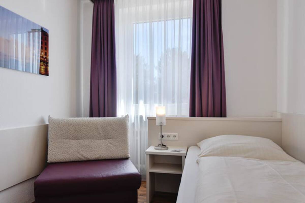 A hotel room with a bed, a nightstand with a lamp, a window with curtains, and a wall-mounted picture.