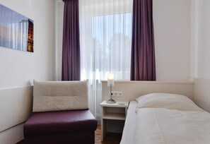 A hotel room with a bed, a nightstand with a lamp, a window with curtains, and a wall-mounted picture. - Hotel Olympia (Munich)