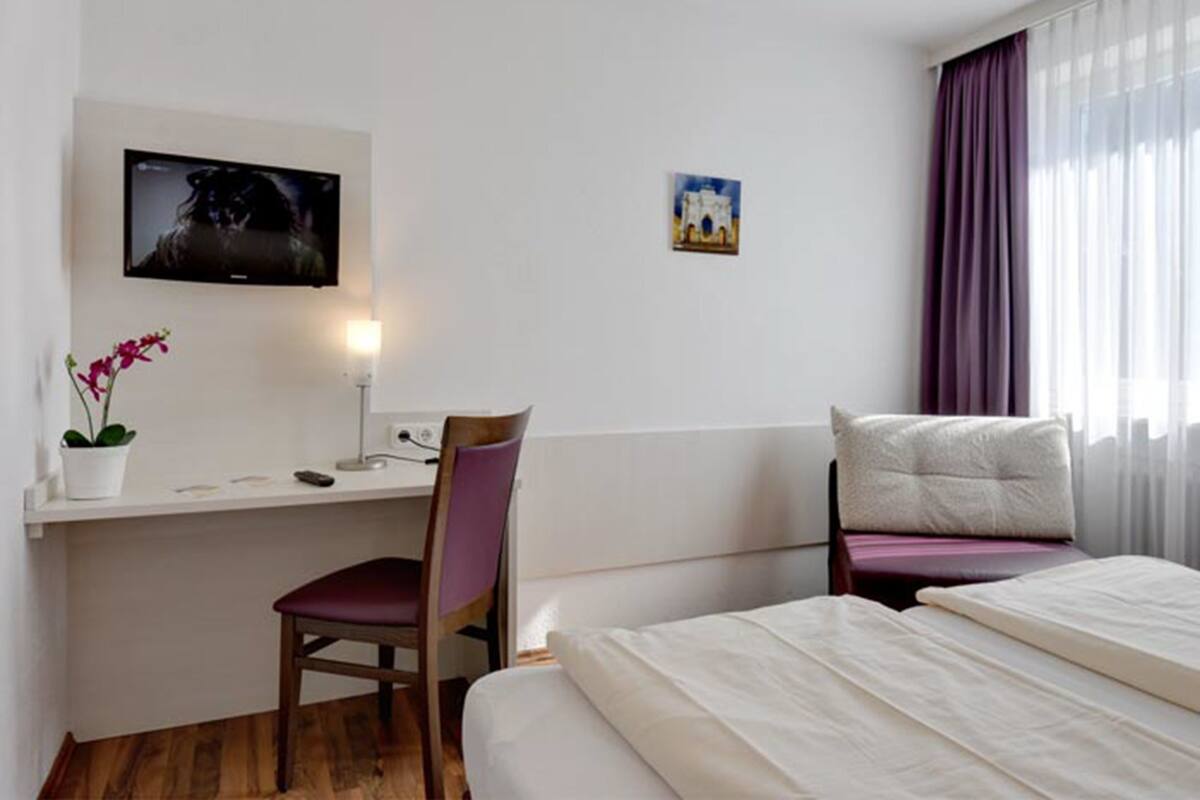A hotel room with a bed, a desk with a chair, a television, a small painting, and a window with curtains.