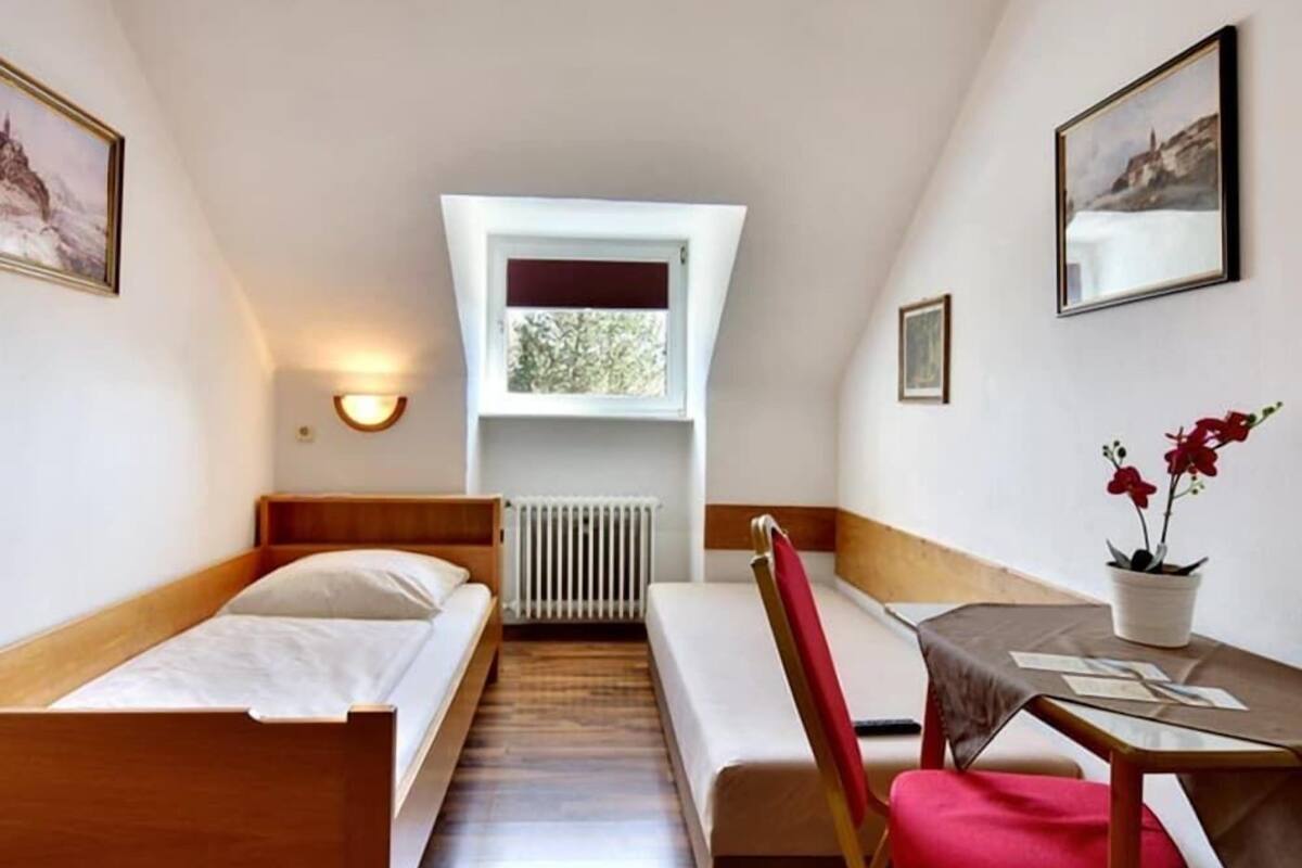 A small, single-room hotel room with a bed, a desk with a chair, a radiator, a window, and a wall-mounted light.