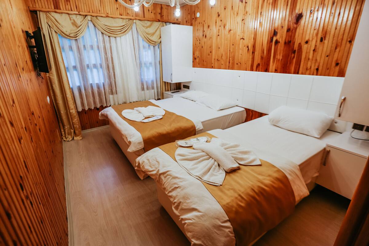 Habitación de hotel con dos camas, paneles de madera, un ventanal con cortinas y una mesita de noche con lámpara.