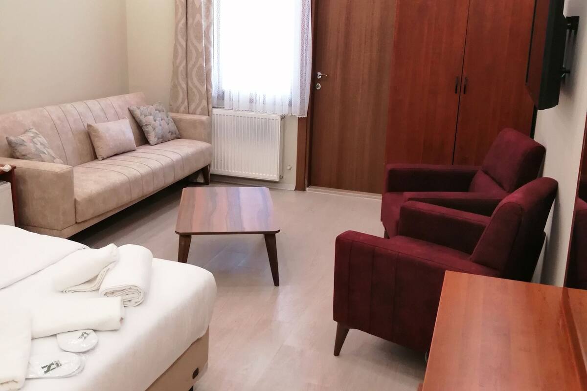 Una habitación luminosa con una cama, un sofá, un sillón rojo, una mesa de madera y un armario.