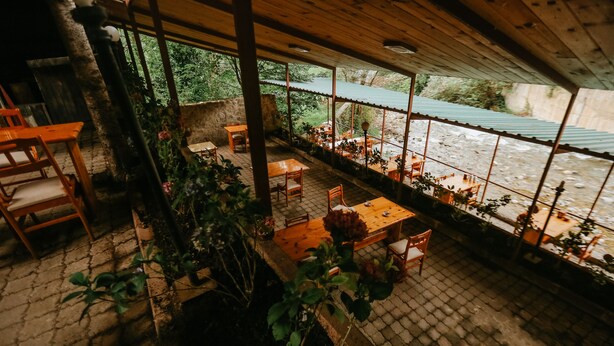 Uma área de refeições ao ar livre com mesas e cadeiras, cercada por plantas e vista para um rio.