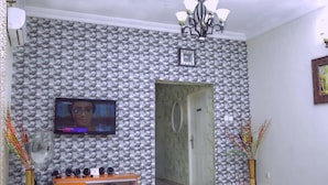 Una sala de estar con un televisor montado en la pared, un mueble de madera con dispositivos electrónicos, una mesa blanca y un papel tapiz estampado.