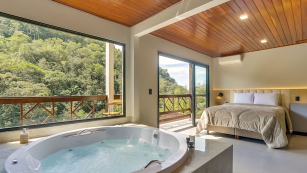 A modern bedroom with a large window, a hot tub, and a bed with a headboard. - Pousada Alpenhof (Sao Pedro de Alcantara)