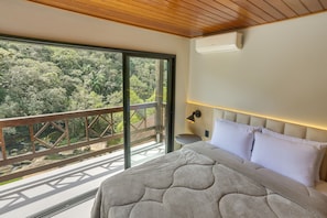 A bedroom with a large bed, a nightstand, a wall-mounted lamp, and a view of a forest through a sliding glass door.