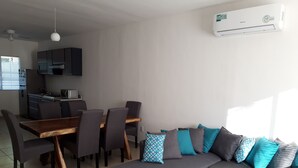 A modern living room with a sofa, a dining table, and chairs. There is a kitchen area in the background with cabinets and a microwave. - Casa Falabella - Puerto Marino (Puerto Morelos)
