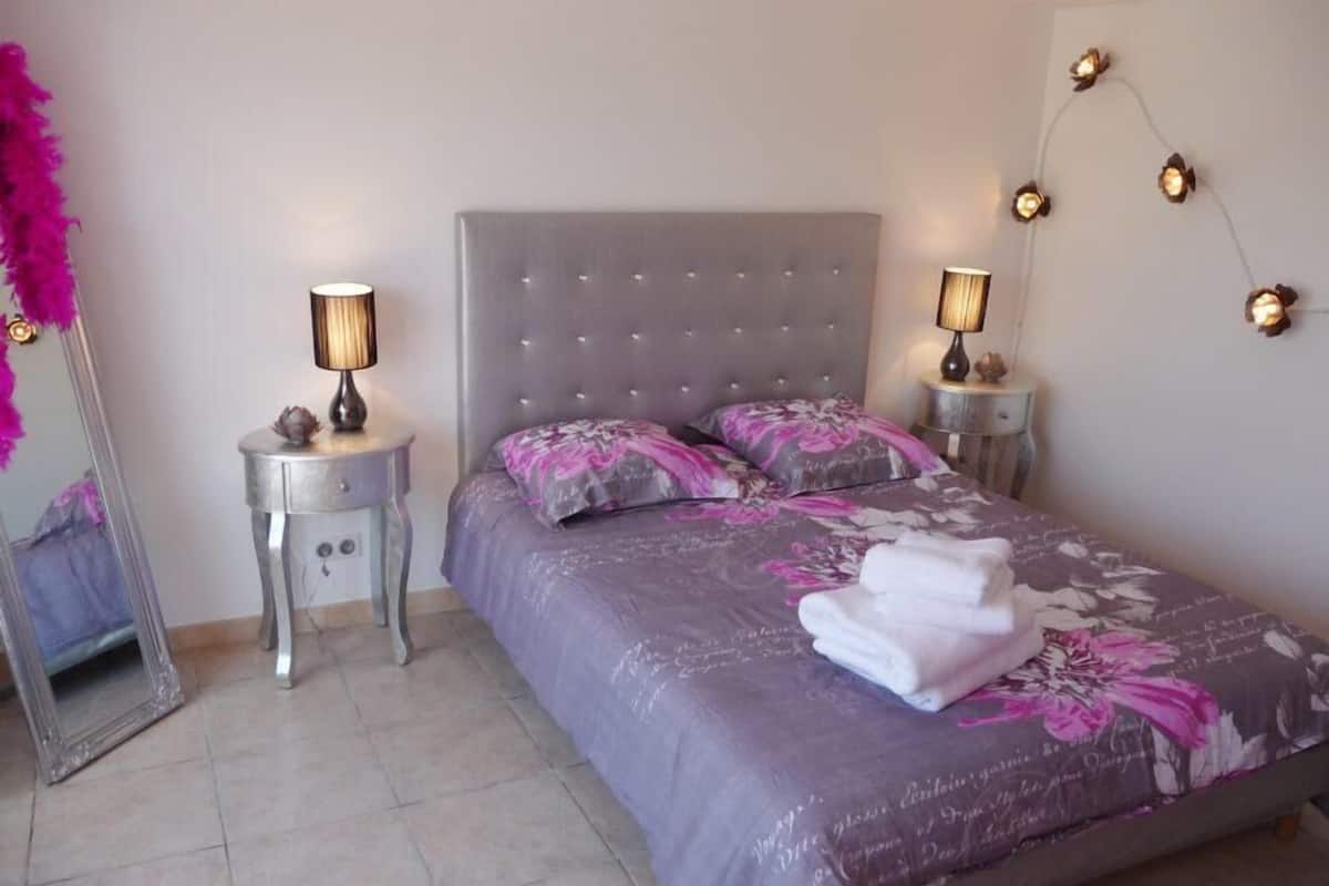 A neatly arranged bedroom with a large bed, two bedside tables with lamps, a mirror, and decorative wall lights.