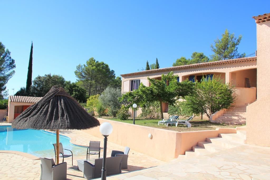 A pool area with lounge chairs and a thatched umbrella, a building with a tiled roof, and a staircase leading to a second floor.