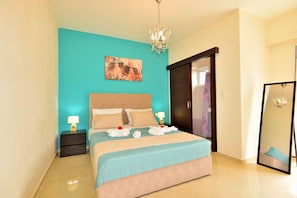 A neatly made bed with a teal bedspread and two pillows, a nightstand with a lamp, a chandelier, and a painting on the wall. - Luxury Suites in Stavromenos (Rethymno)