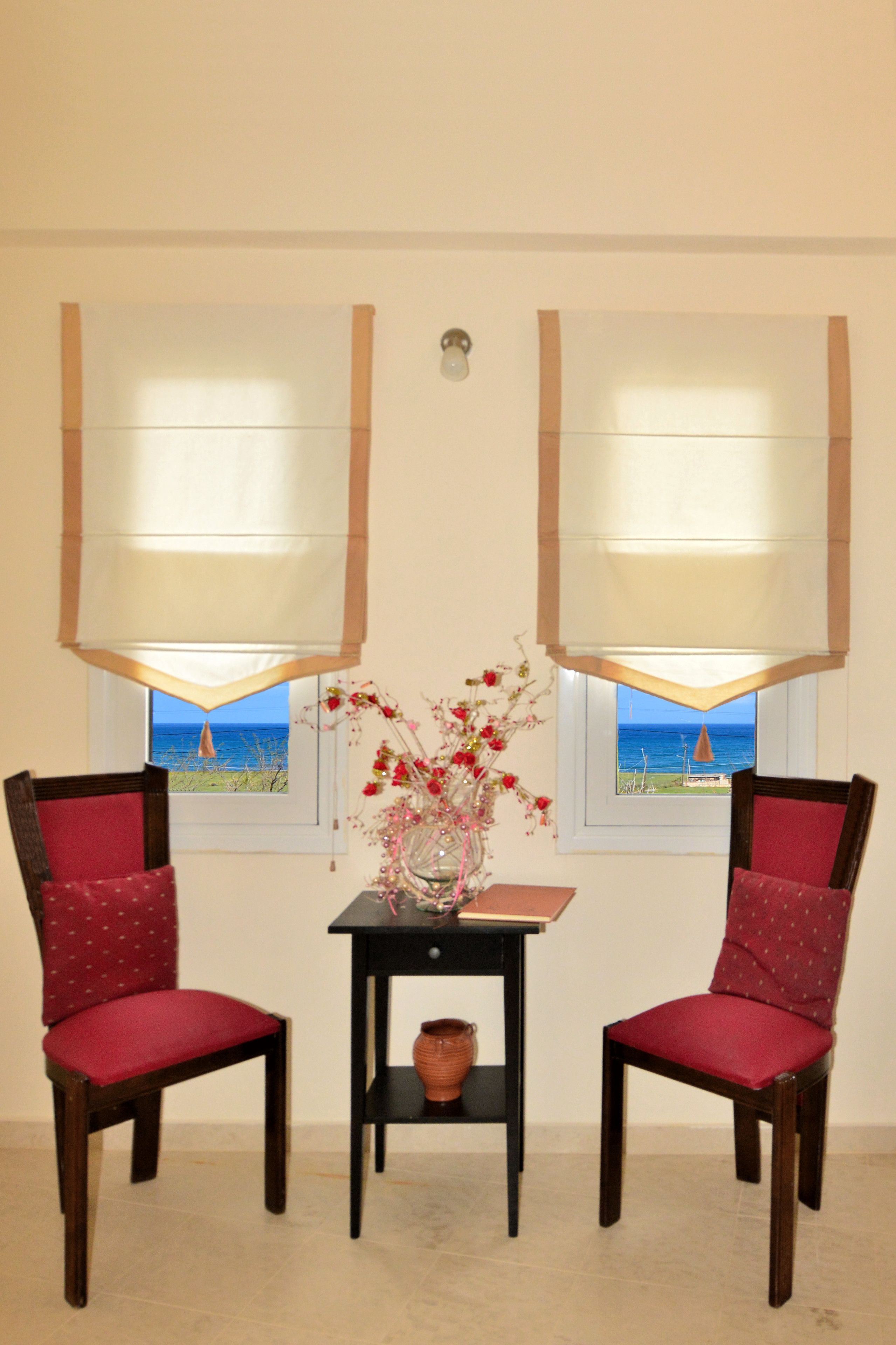A room with two chairs, a small table, and a vase with flowers. There are windows with blinds, and a view of the sea is visible through the windows.