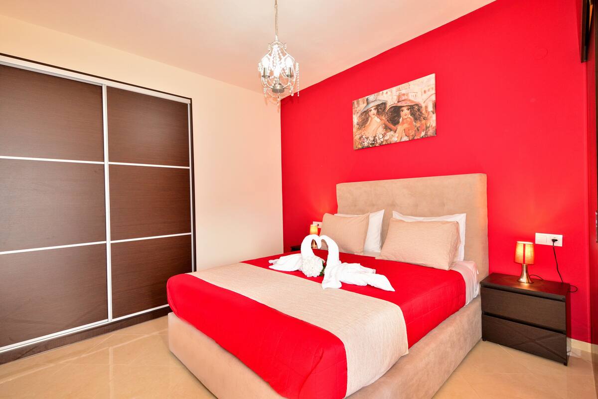 A hotel room with a large bed, a red wall, a chandelier, and a painting.