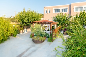 A courtyard with a wooden pergola, potted plants, and a building with a beige facade. - Luxury Suites in Stavromenos (Rethymno)
