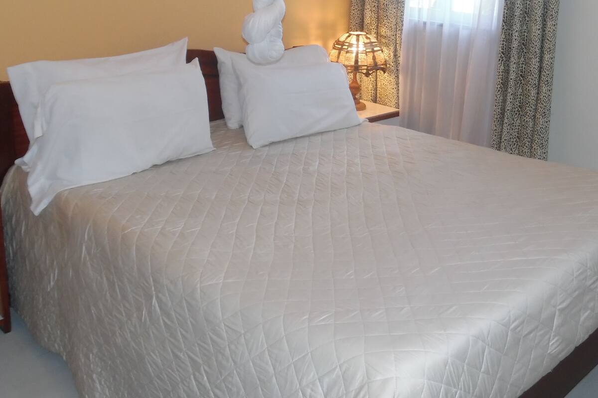 A neatly made bed with a quilted white duvet and two pillows, a bedside lamp, and a curtain with a leopard print pattern.