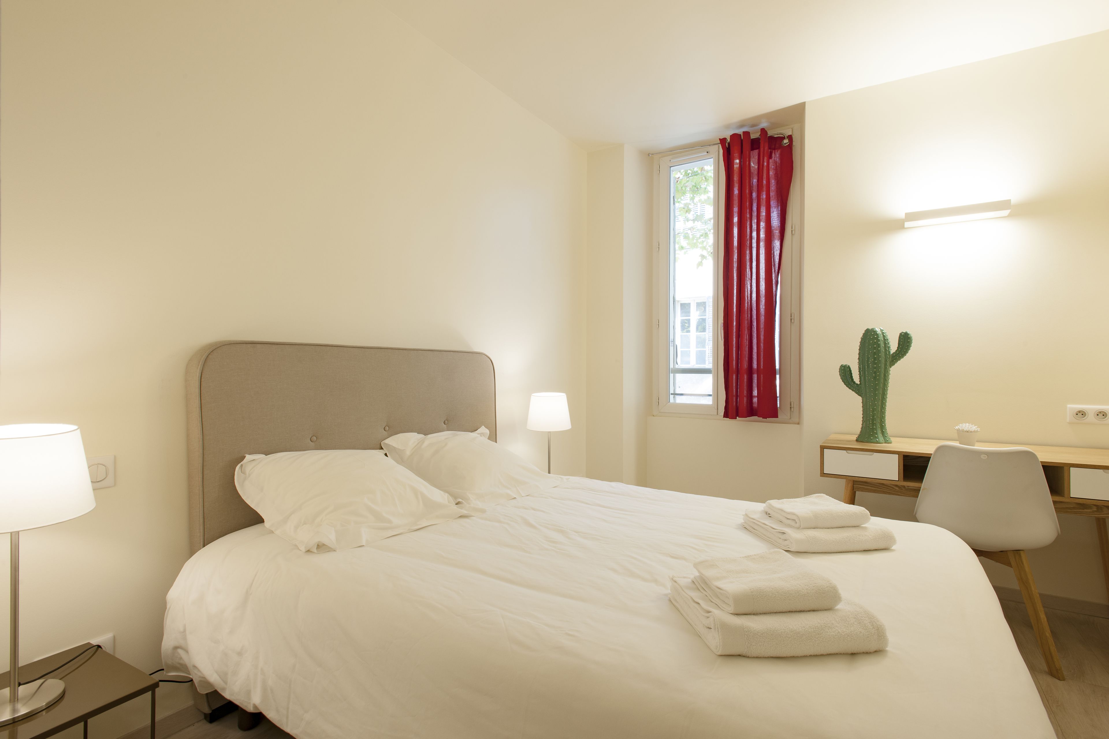 Un lit bien fait, avec du linge de lit blanc et des serviettes pliées, un bureau avec une chaise, un cactus et une fenêtre avec des rideaux rouges.