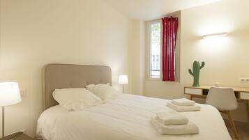 A neatly made bed with white linens and folded towels, a desk with a chair, a cactus, and a window with red curtains.