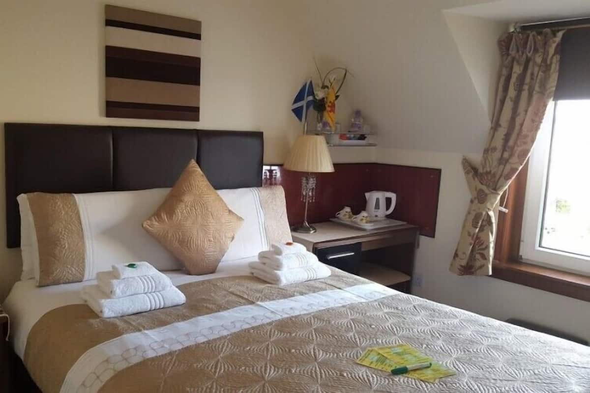 Une chambre d’hôtel avec un grand lit, une table de chevet avec une lampe, un petit bureau avec une bouilloire et une fenêtre avec des rideaux.