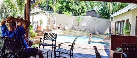A covered outdoor area with a pool, wooden deck, and chairs.