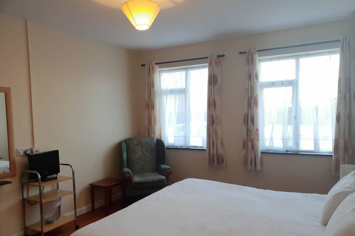 A hotel room with a bed, a chair, a desk with a computer, and a window with curtains.