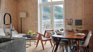 A wooden interior with a dining area, a sofa, a table with chairs, a lamp, and a view of the sea through large windows. - Nusfjord Village & Resort (Flakstad)