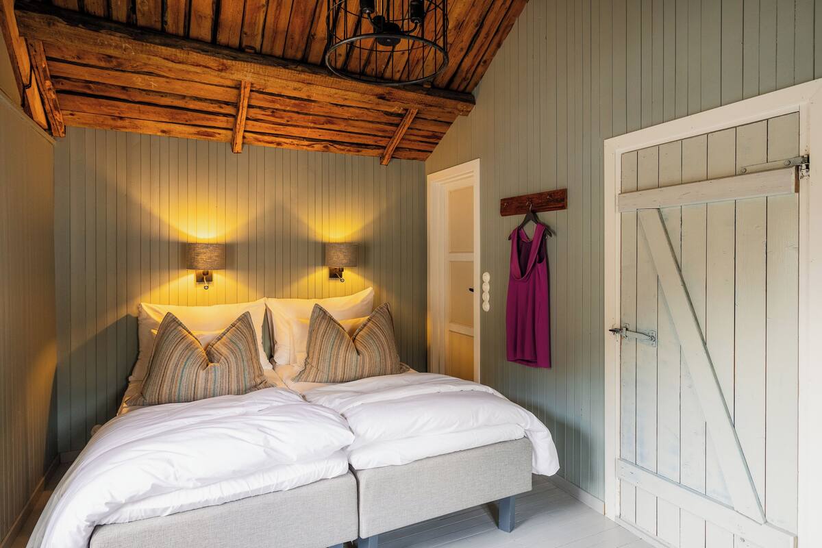 Un dormitorio con una cama, dos lámparas adosadas a la pared, techo de madera y una puerta blanca.