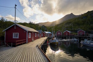 Un muelle de madera que lleva a edificios rojos, barcos amarrados y un paisaje montañoso al fondo.