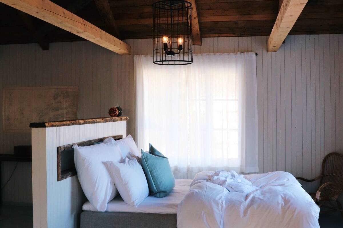 Un dormitorio con una cama, cabecera de madera, ventana con cortinas y una lámpara colgante.