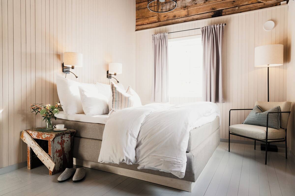 Un dormitorio con cama, lámparas de noche, una silla, una mesita con un florero y una ventana con cortinas.
