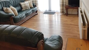A living room with a green sofa, a wooden coffee table, and a television. There is a window with curtains and a door leading outside. - Holly Cottage (Skegness)
