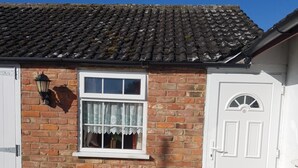 A small brick house with a tiled roof, a white door, and a window with a white frame and lace curtain. - Holly Cottage (Skegness)