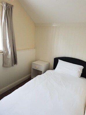 A single bed with white bedding, a small bedside table with a drawer, and a window with curtains.