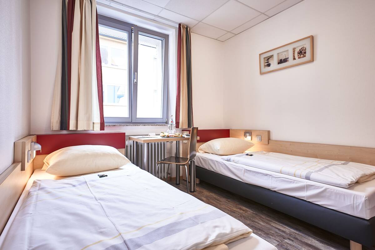 Habitación de hotel con dos camas individuales, una silla, una mesita con hervidor y una ventana con cortinas.