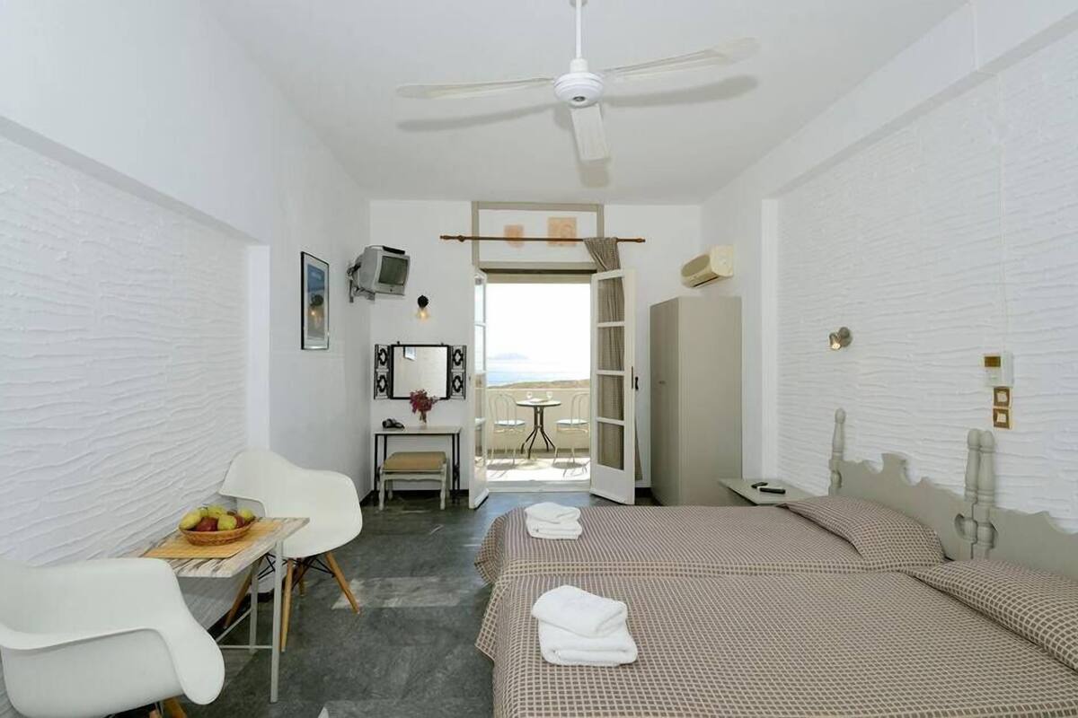 A hotel room with two beds, a desk, a chair, a small table with fruit, a ceiling fan, and a view through an open door.