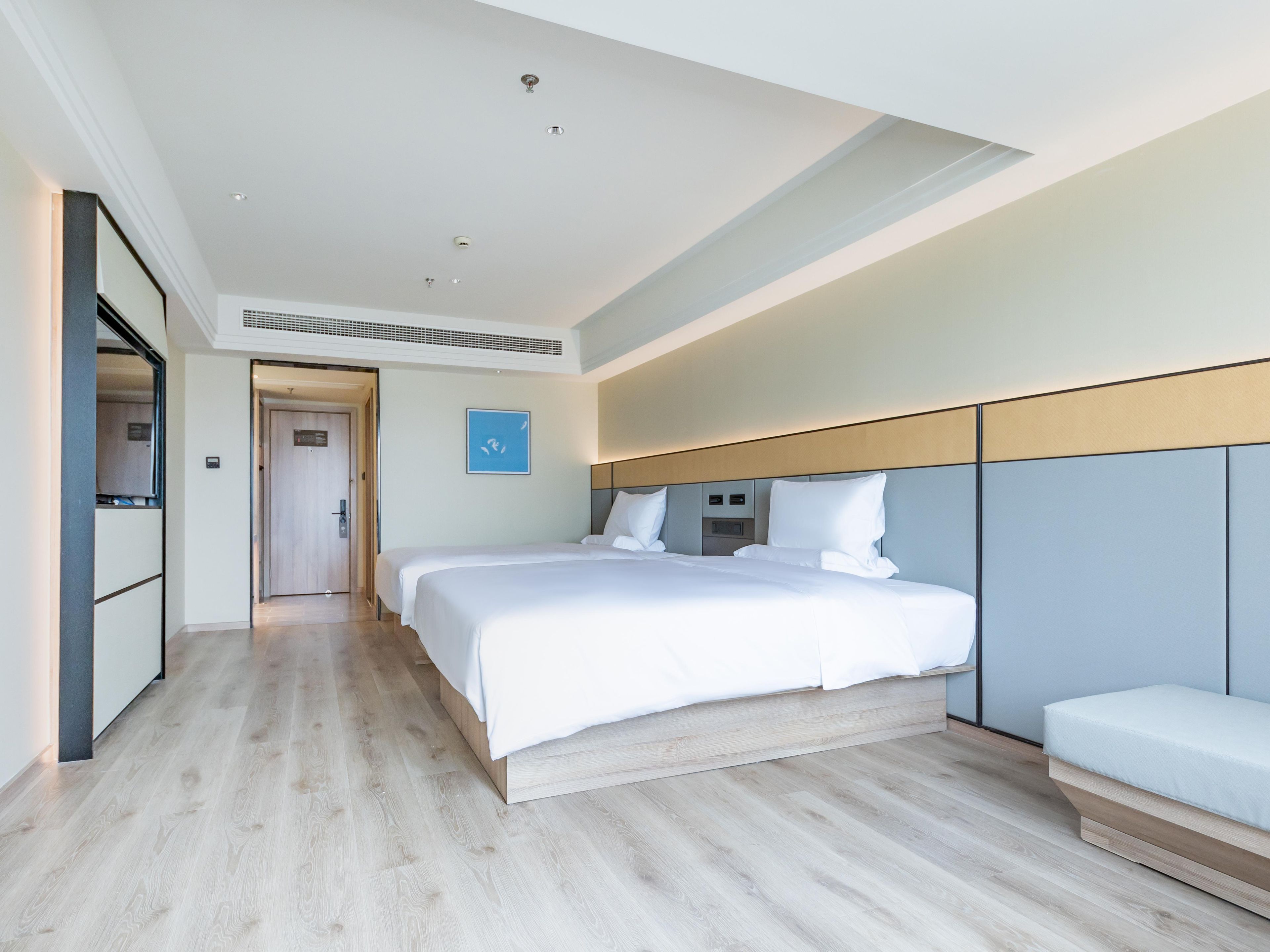 A modern hotel room with a large bed, a bedside table, a built-in bench, and a view of the hallway.