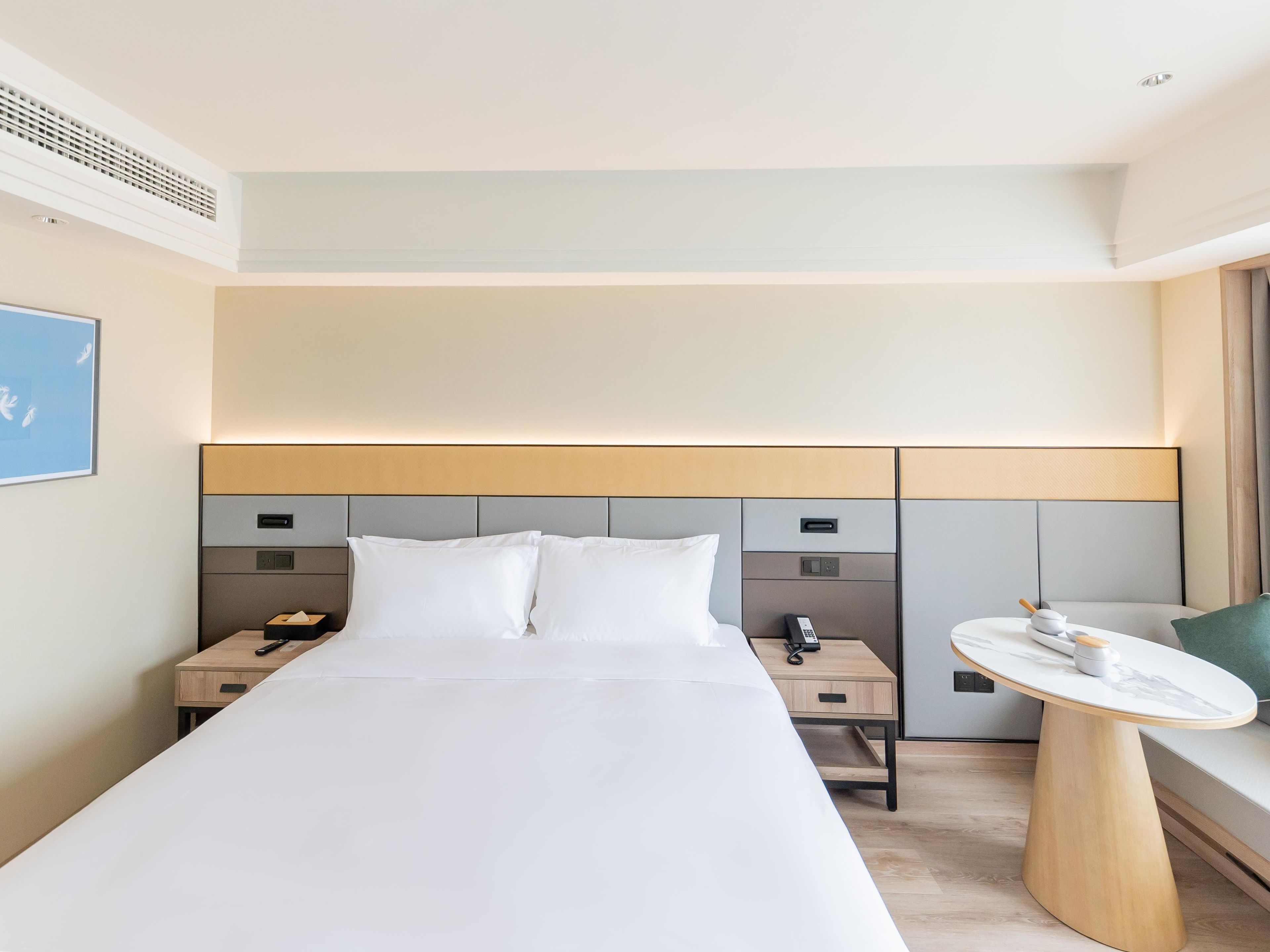 A modern hotel room with a large bed, bedside tables, a small round table, and a headboard with built-in controls.