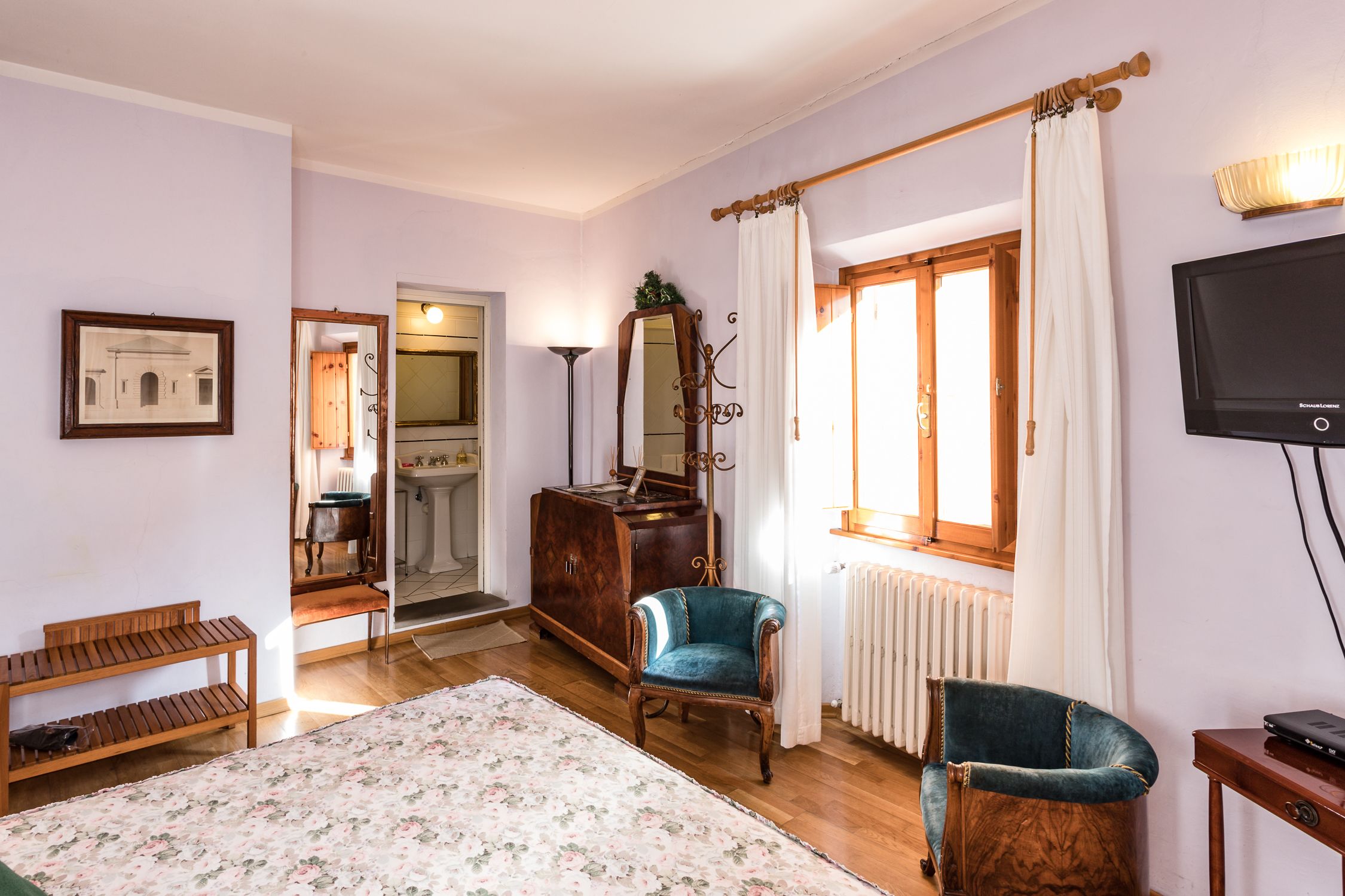 A room with a bed, a wooden bench, a blue armchair, a dresser, a mirror, a radiator, and a TV.