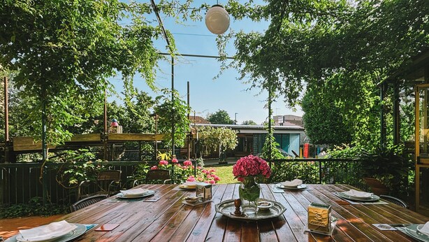An outdoor dining area with wooden tables set for two, surrounded by lush greenery and a view of a building in the background. - Hotel la Felicina Mugello (Scarperia e San Piero)