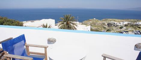 Una terraza con una mesa y sillas blancas, con vistas a un paisaje costero con edificios y al mar.