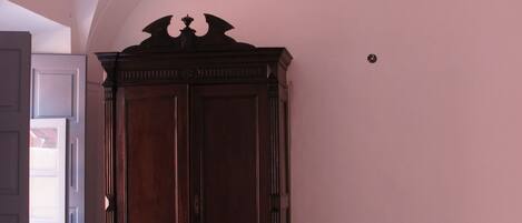 A room with a wooden wardrobe, a chair, two beds, and a table.