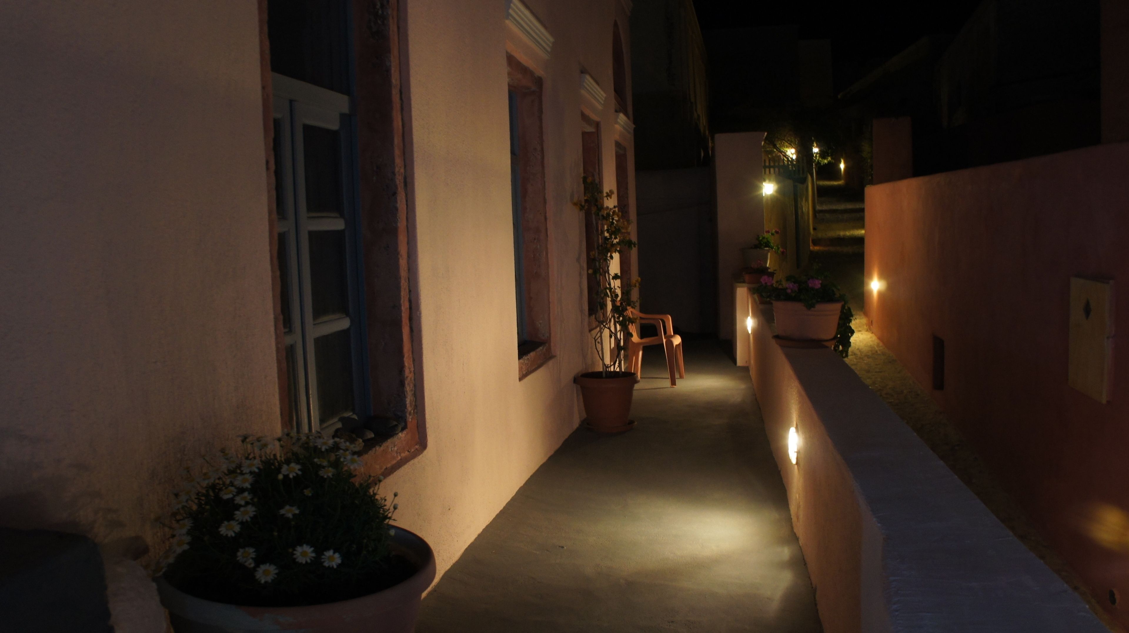 A narrow alleyway with potted plants and a chair, illuminated by wall-mounted lights.
