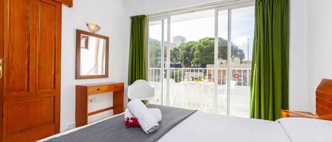 Una camera d'albergo con un letto, una scrivania, una sedia, uno specchio e un balcone con tende verdi.