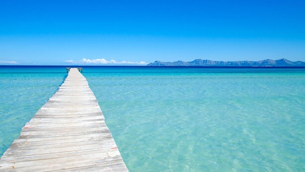 A wooden pier extending into clear turquoise water with a mountain range in the distance.