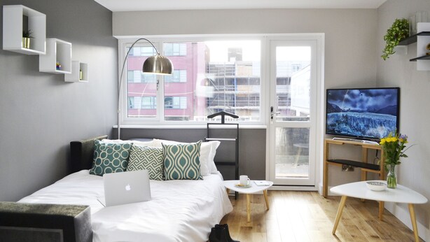 Quarto moderno com cama, laptop, TV e vista para edifícios pela janela.