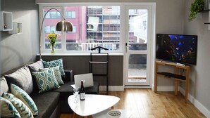 Uma sala de estar moderna com um sofá cinzento, uma mesa de centro branca, uma televisão sobre um suporte de madeira, e uma janela com vista para edifícios.