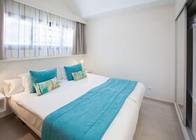 Een net opgemaakt bed met een blauwe deken en meerdere kussens, een nachtkastje en een raam met gordijnen.